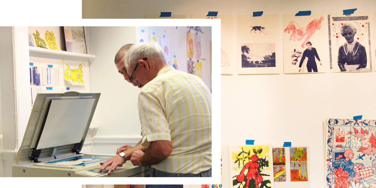 Tommy showing David how to set up the machine alongside a wall of students work