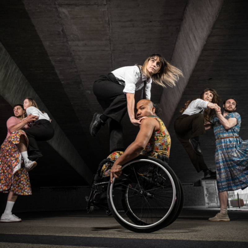 A photograph taken underneath an underpass of a group of six Stopgap dancers. The dancers are wearing either colourful patterned dress or black trousers with white shirts.