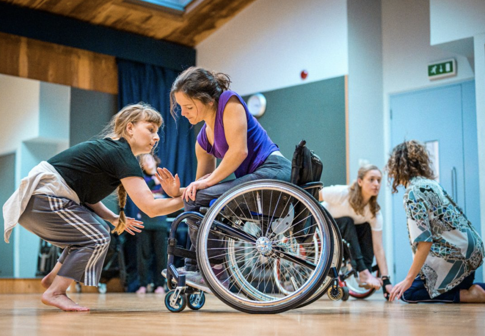 A studio shot of a dancer in a wheelchair opposite an able bodied dancer with their arms intertwined. In the background other dancers are interacting with one another in the studio space.