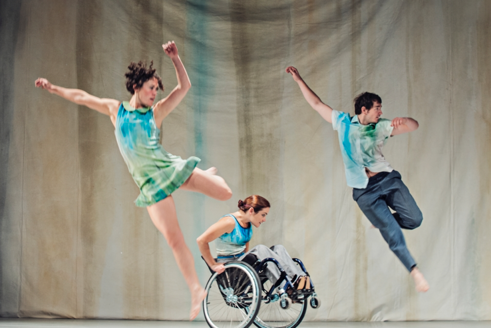 Three dancers performing on stage, two of the dancers are captured mid movement leaping into the air whilst the third dancer is balancing in a wheelie position in their wheelchair.